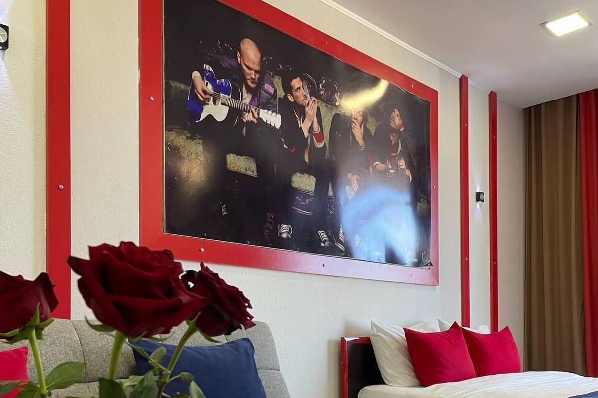 A hotel room with a grey sofa, a bed with red and white bedding, a large poster on the wall, and a vase with roses on the table.