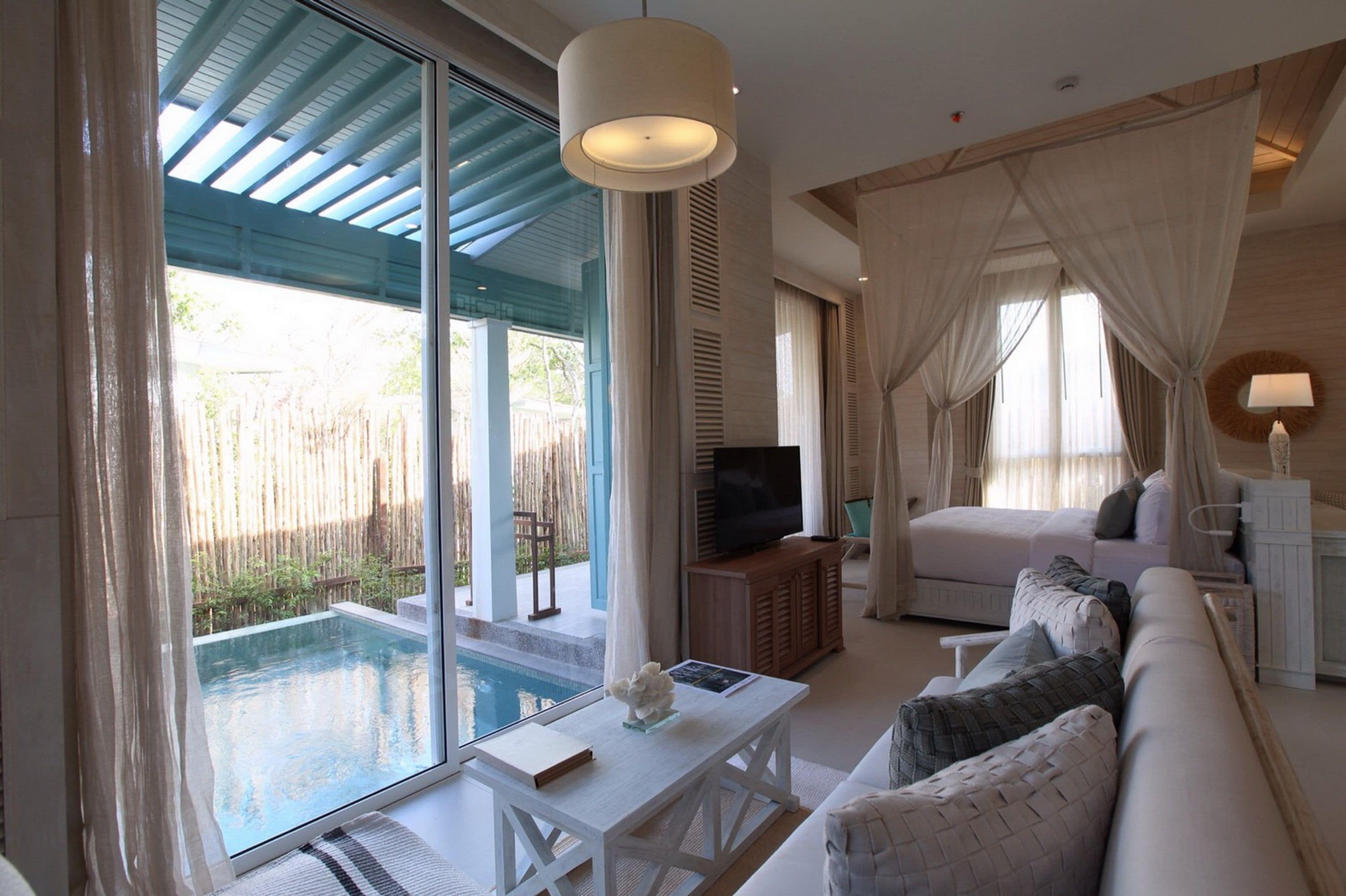 A modern hotel room with a large glass door leading to a pool, a sofa, a TV, and a bedside table.