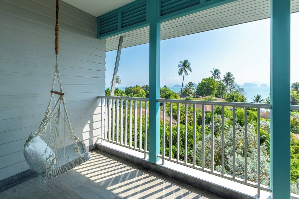 Un balcon agrémenté d’un hamac suspendu, de balustrades blanches et offrant une vue sur un paysage tropical, avec des palmiers et une étendue d’eau.