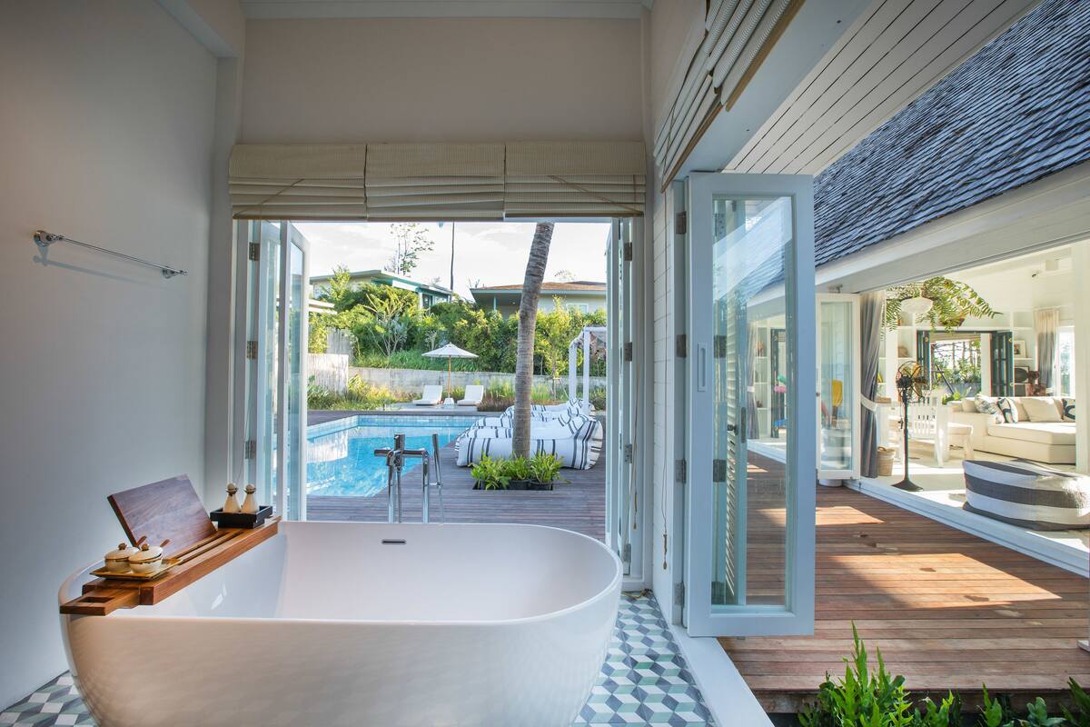 Une salle de bain moderne avec une baignoire sur pieds, une étagère en bois agrémentée d’objets décoratifs, et une vue sur une piscine extérieure et sur du mobilier de jardin.
