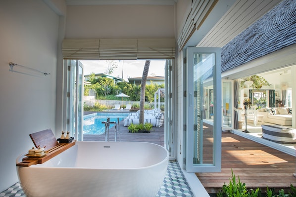 Une salle de bain moderne avec une baignoire sur pieds, une étagère en bois agrémentée d’objets décoratifs, et une vue sur une piscine extérieure et sur du mobilier de jardin.