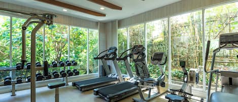 A gym with treadmills, ellipticals, and dumbbells, large windows with a view of greenery.