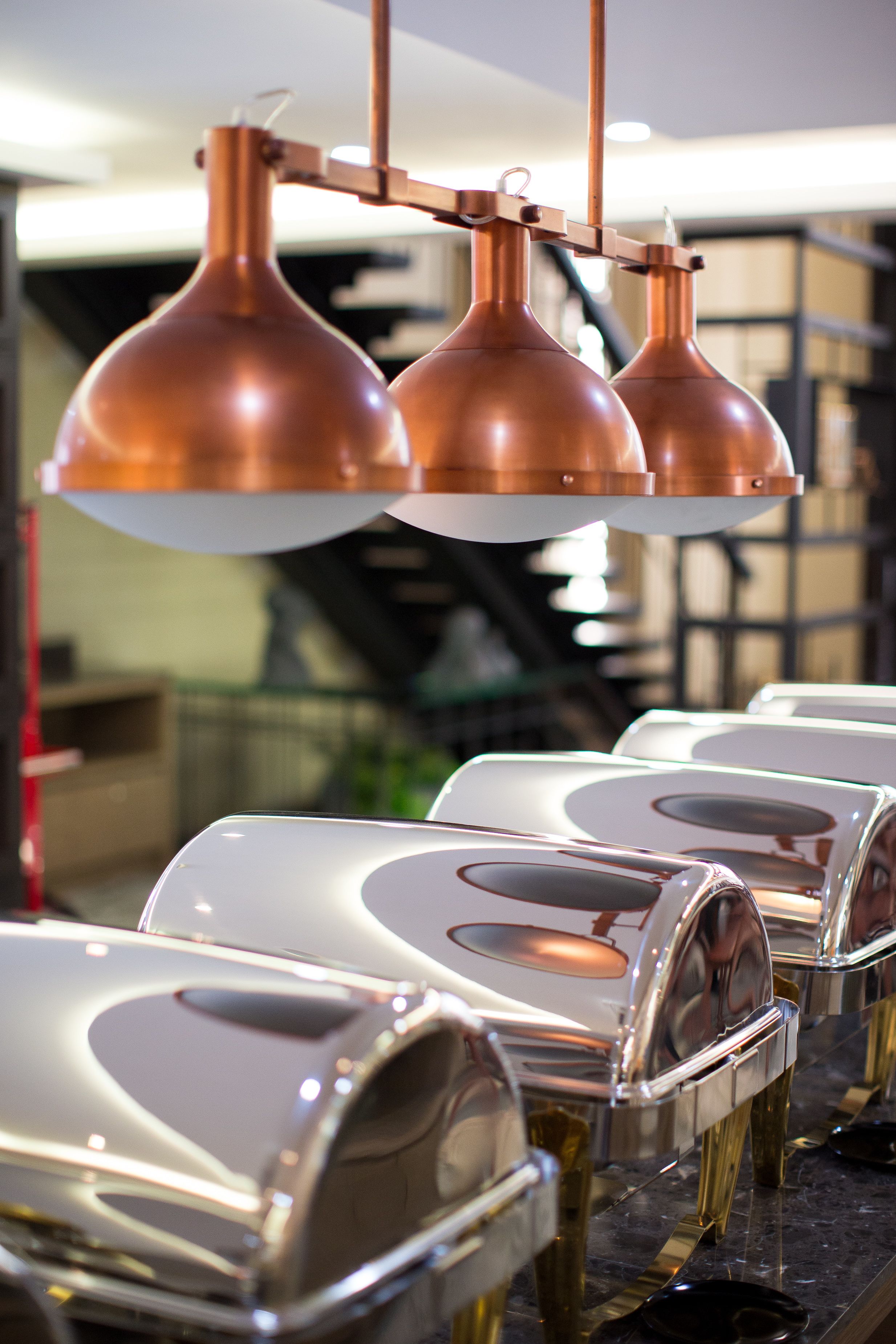 A row of reflective, metallic serving dishes and copper pendant lights in a commercial kitchen setting.