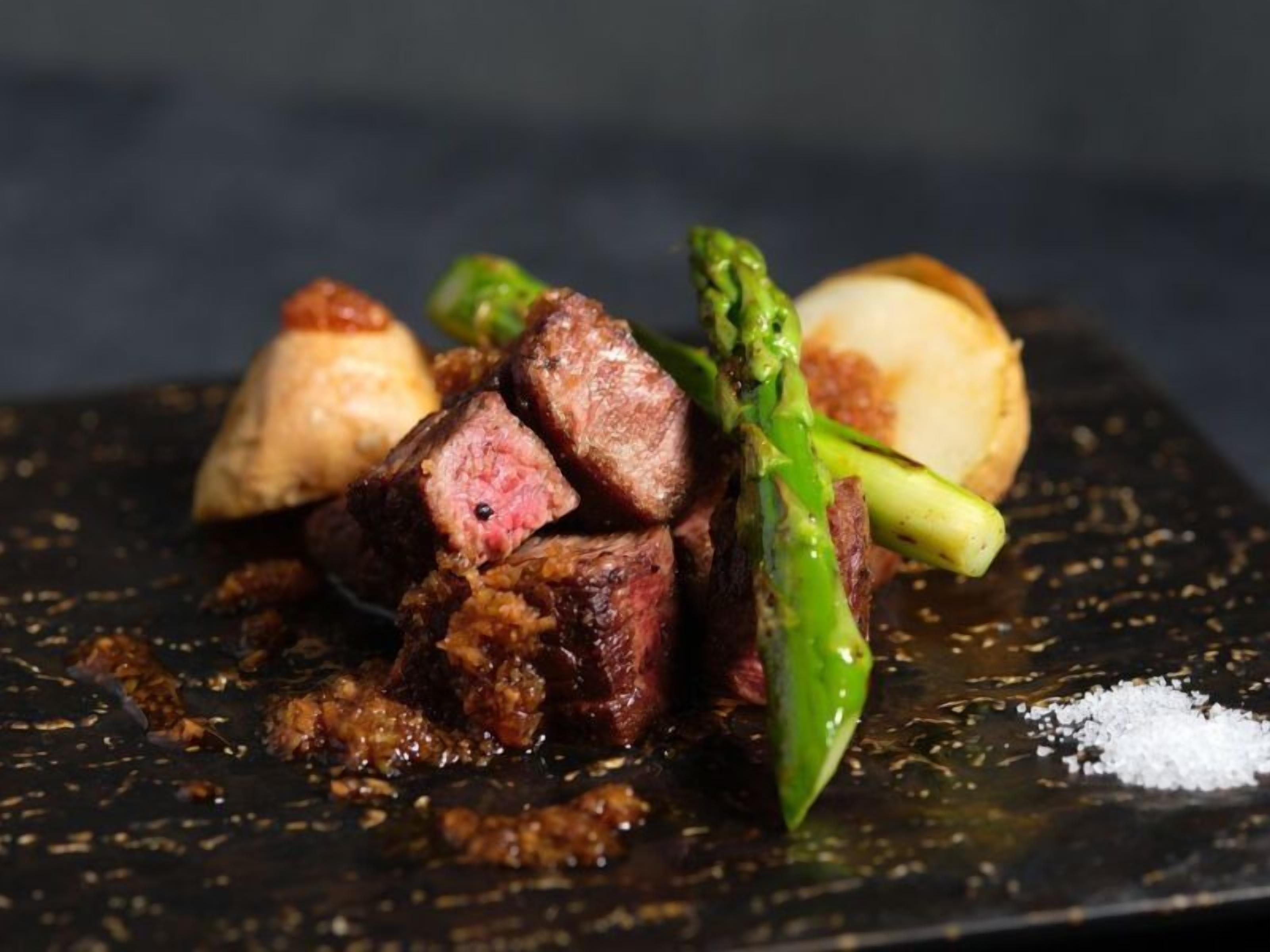 A dish with steak, asparagus, and a round potato on a dark plate.