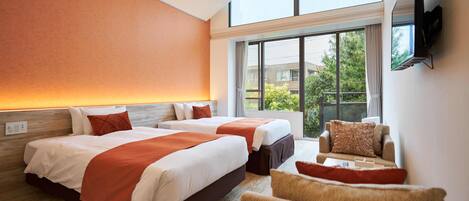 A hotel room with two beds, a sofa, a TV mounted on the wall, and a large window offering a view of greenery outside.