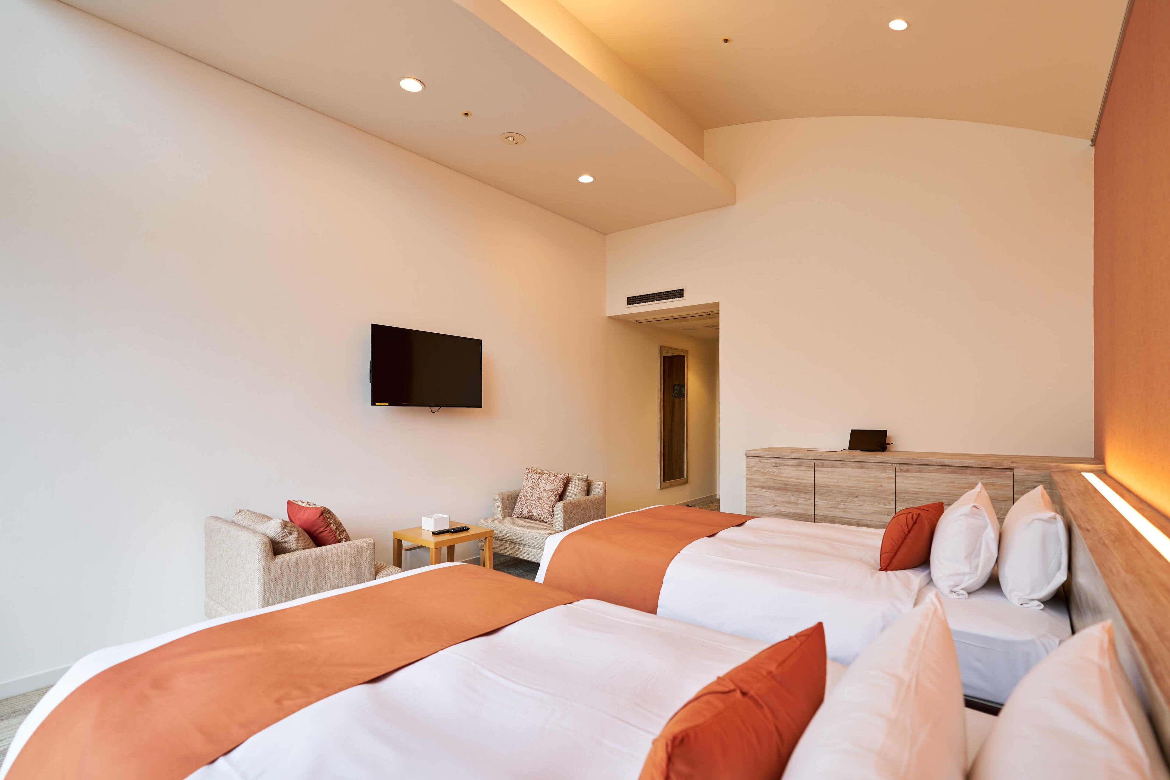 A hotel room with two beds, a TV mounted on the wall, a small table, and a sofa.