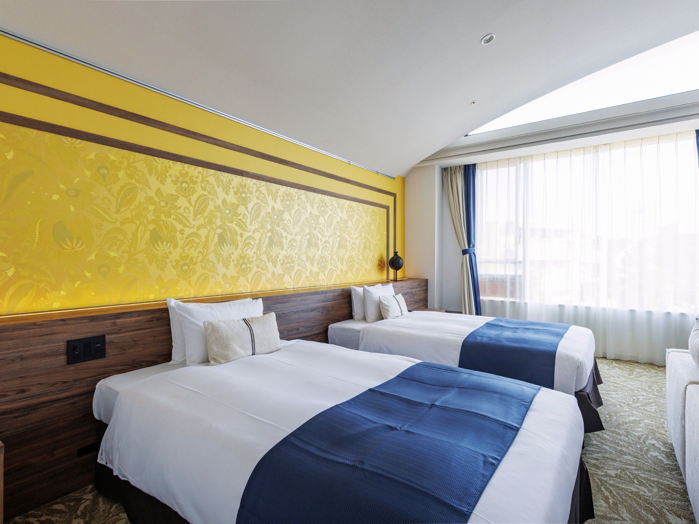 A hotel room with two beds, a wooden headboard, a patterned wall, and a large window with curtains.