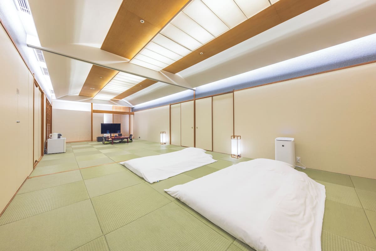 Een traditionele Japanse kamer met tatami vloeren, een laag plafond met houten balken en schuifdeuren. Er staan twee bedden met witte beddengoed, een televisie en een klein tafeltje met een lamp.