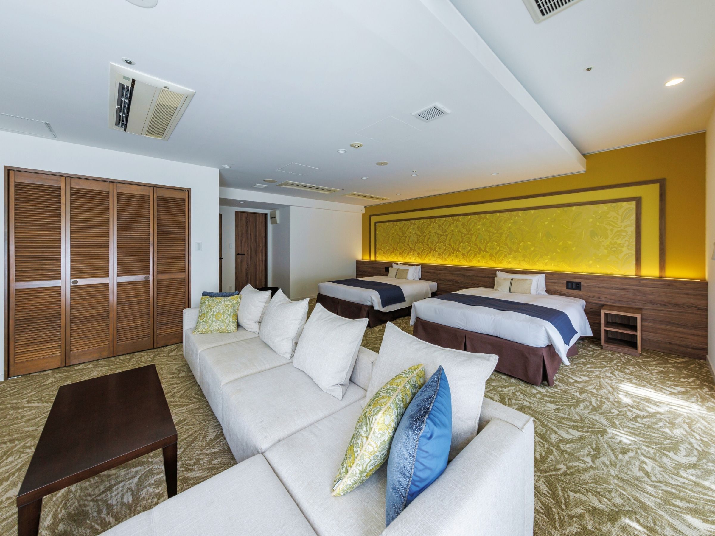 A hotel room with a sofa, two single beds, a wooden wardrobe, and a patterned carpet.