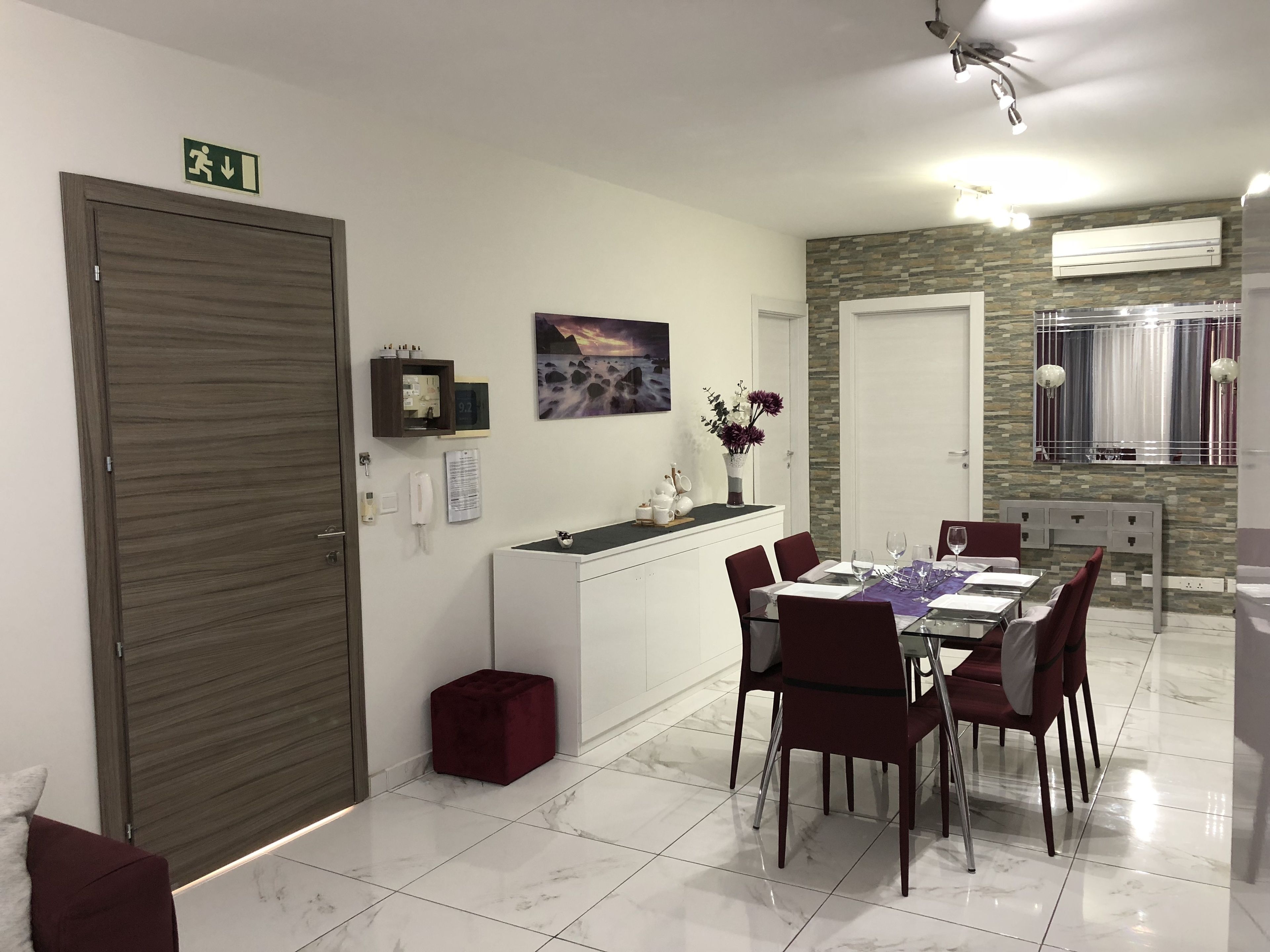 Un coin repas avec un sol en marbre, une table blanche avec des chaises rouges, un petit tabouret rouge, un téléphone fixé au mur, un tableau encadré et une horloge.