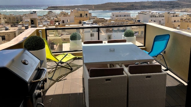 A balcony with a table and chairs, a grill, and potted plants. - Marsascala Sea View Luxury Apartment & Penthouse (Marsaskala)
