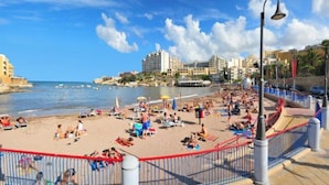 A beach with people sunbathing and swimming, a promenade with a railing, and buildings in the background. - Marsascala Sea View Luxury Apartment & Penthouse (Marsaskala)