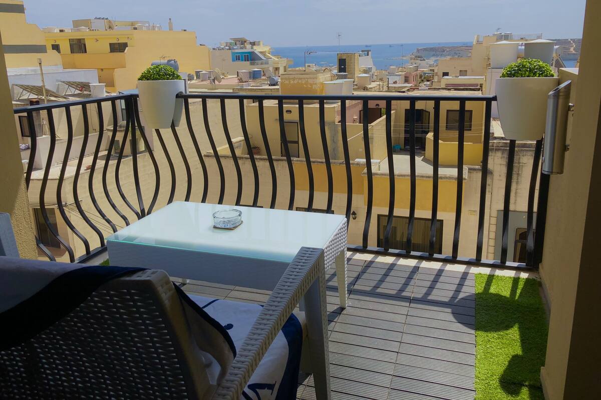 A balcony with a table and chairs, a view of the sea, and buildings in the background.