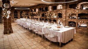A long dining table set with white tablecloths, clear chairs, and wine bottles in a brick-walled room with a fireplace. - Hotel Dolphin (Senec)