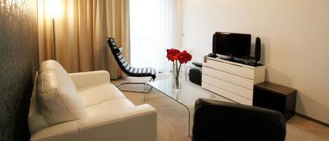 A modern hotel room with a white sofa, a black chair, a glass coffee table, a flat-screen TV on a white cabinet, and a vase with red flowers.