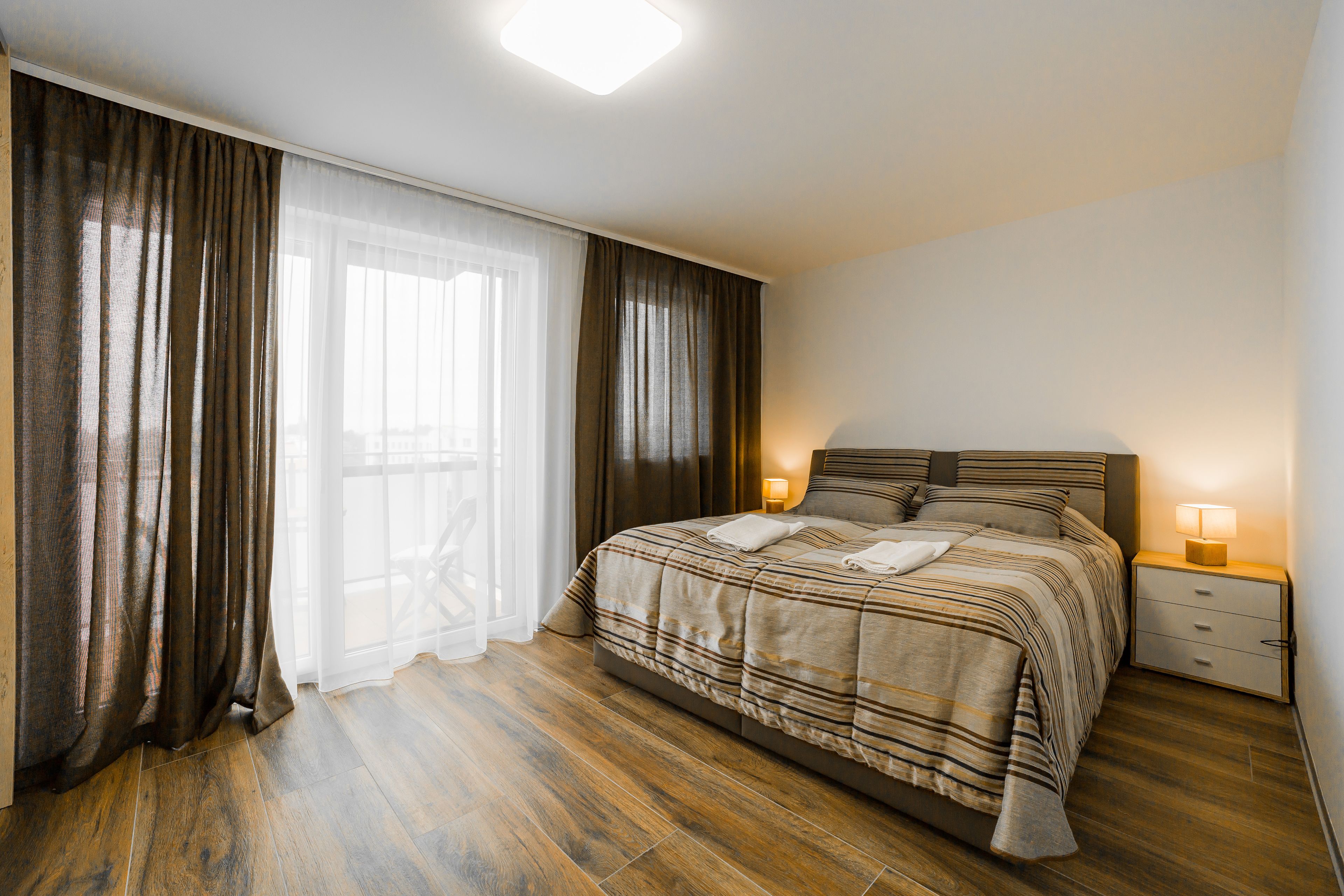 A bedroom with a bed, bedside lamps, a nightstand, and a view through a window with curtains.
