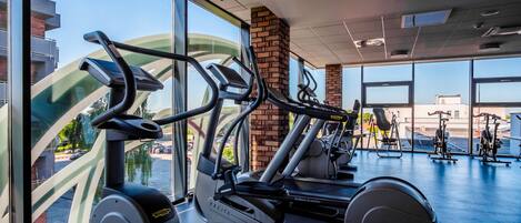 Un gimnasio con máquinas de cardio, un pilar de ladrillo y grandes ventanales con vistas al exterior.