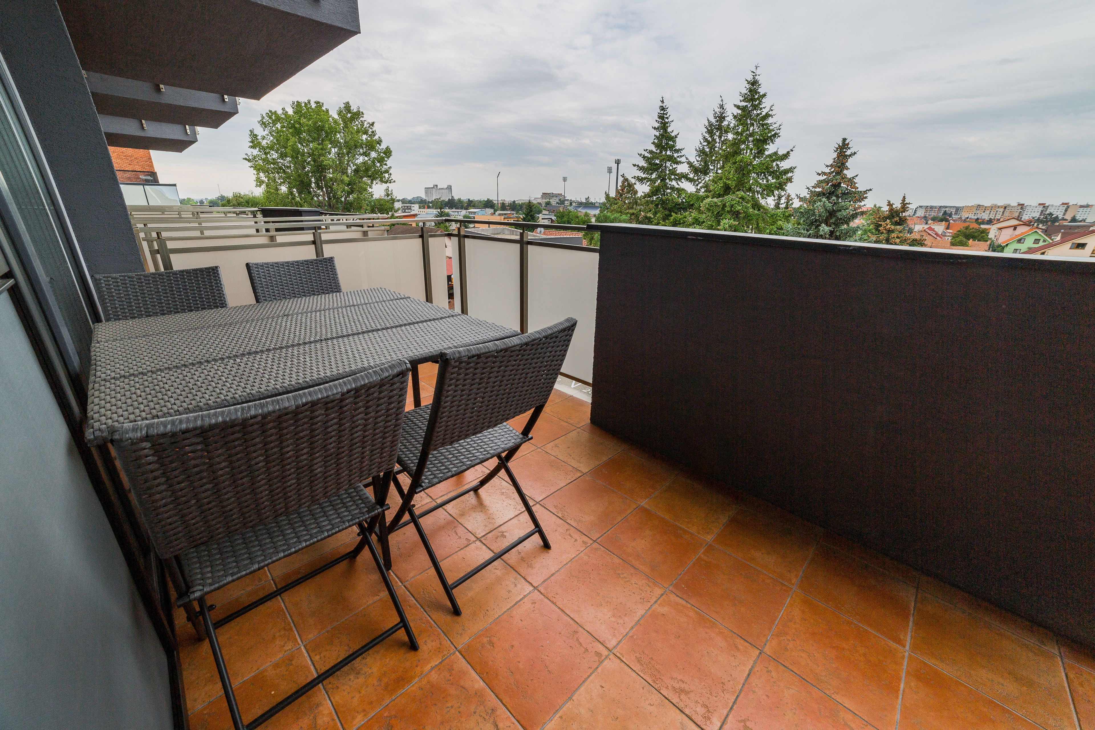 A balcony with a table and chairs, a tiled floor, and a view of buildings and trees.
