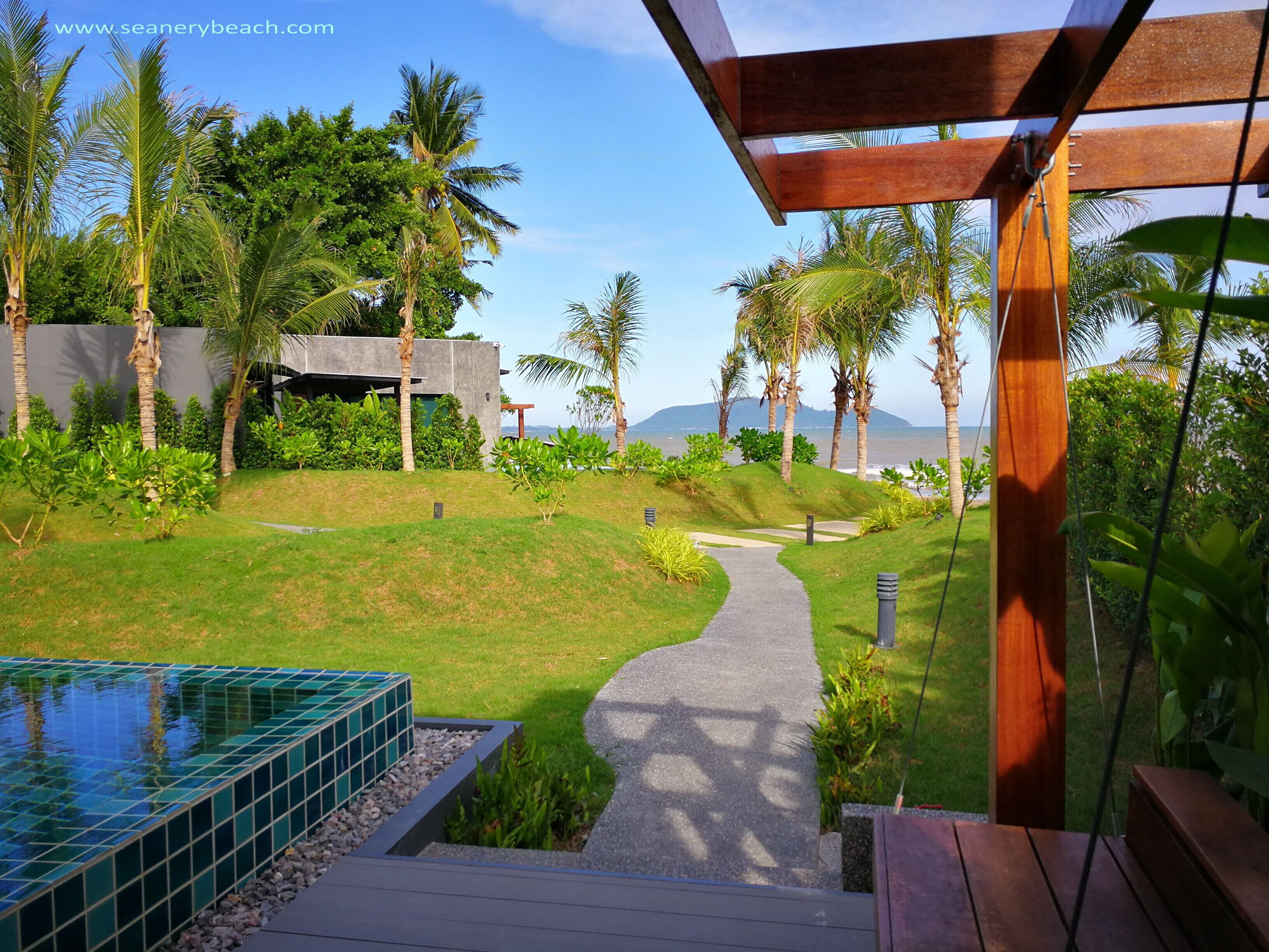 A pathway leading to a pool surrounded by lush greenery and palm trees, with a modern building in the background.
