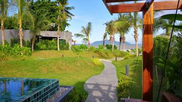 A pathway leading to a pool surrounded by lush greenery and palm trees, with a modern building in the background.