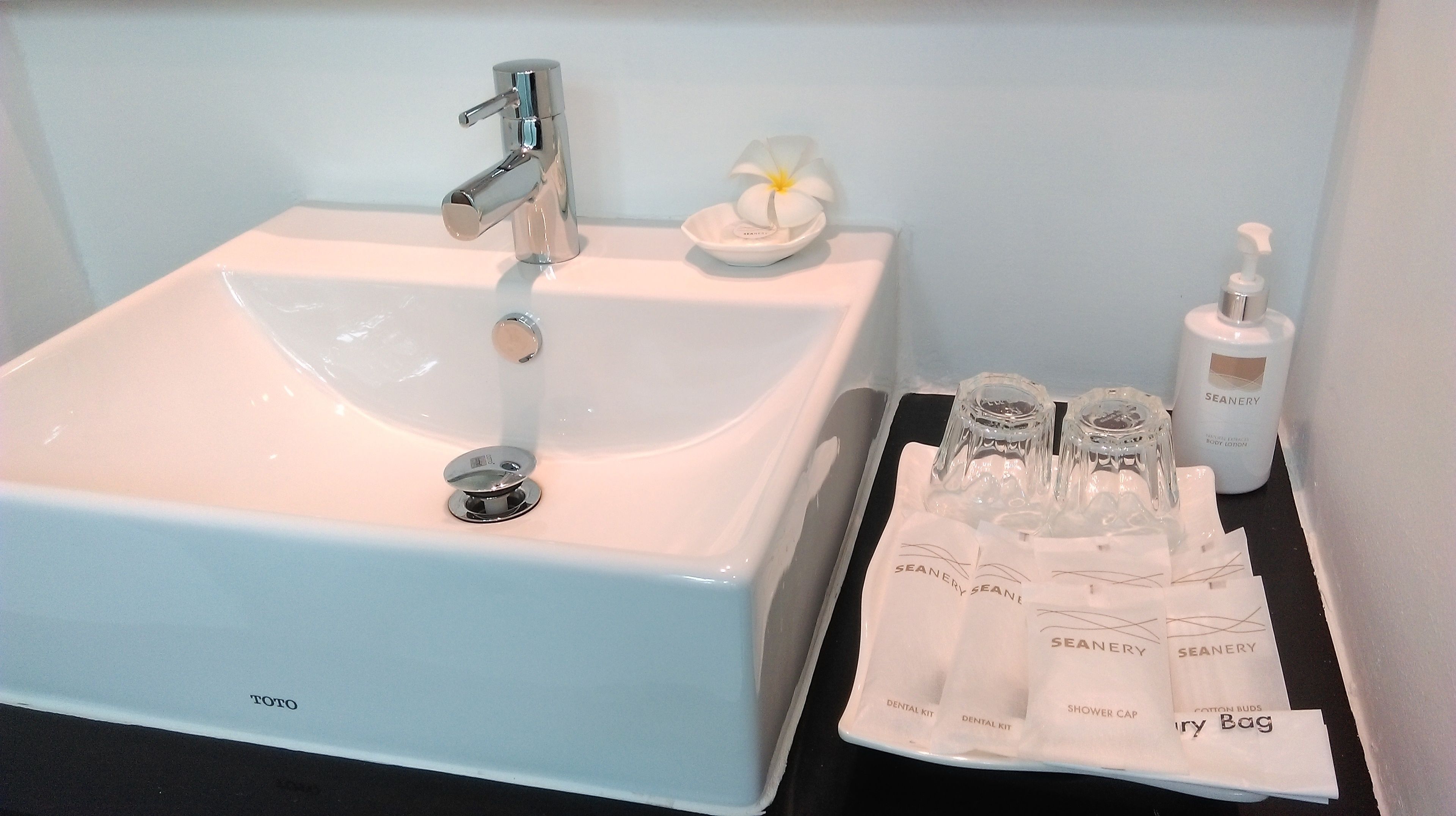 A bathroom with a white sink, a mirror, and toiletries including a bottle of Seanery body lotion, a dental kit, and a shower cap.