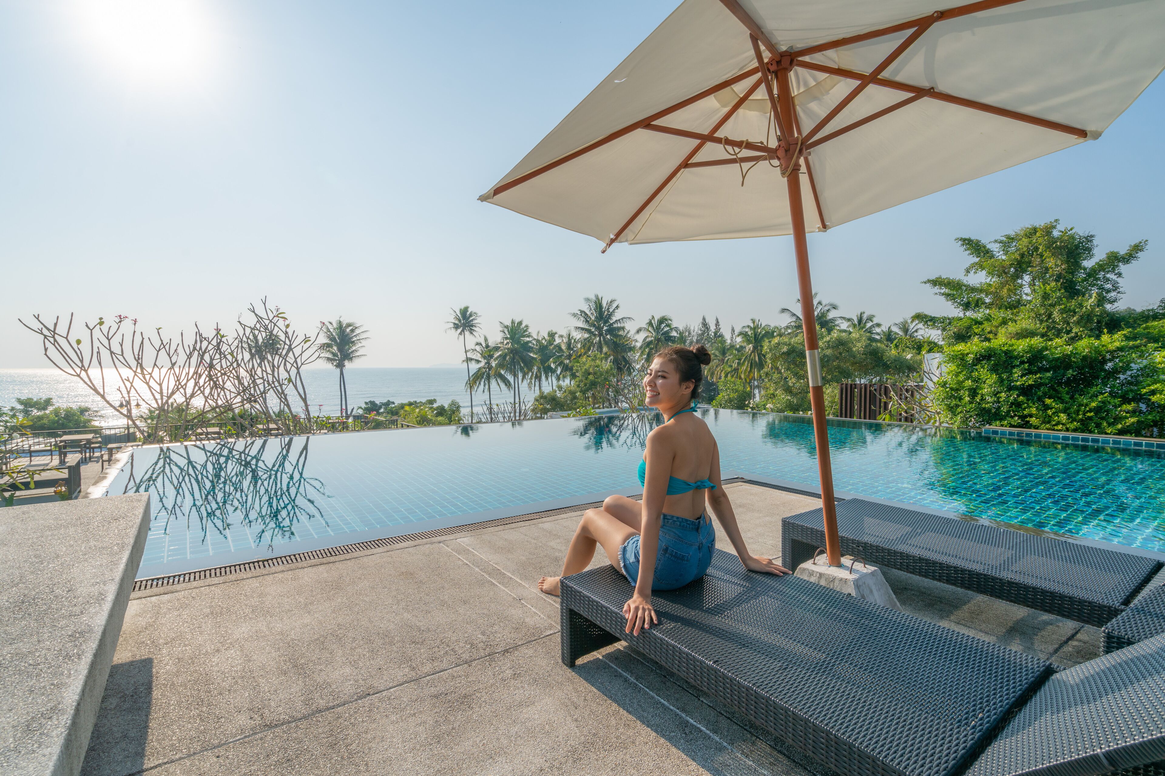 A person sitting on a lounge chair by a pool with a view of the sea and palm trees.