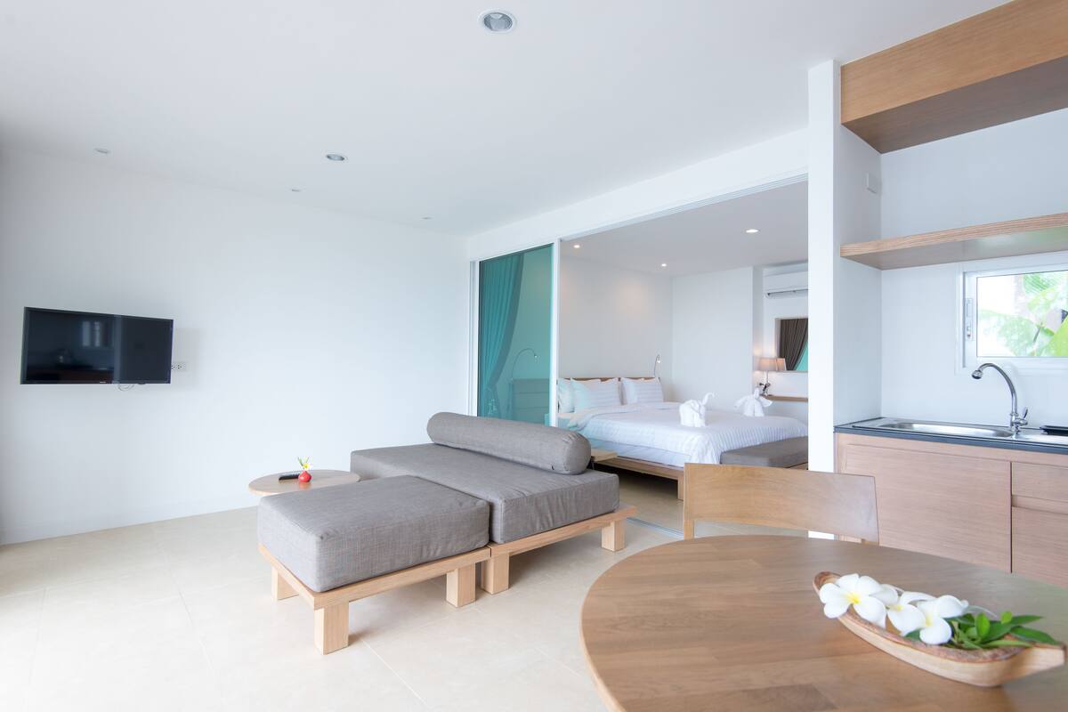 A modern hotel room with a sofa bed, a dining table with a bowl of flowers, a TV, and a kitchenette.