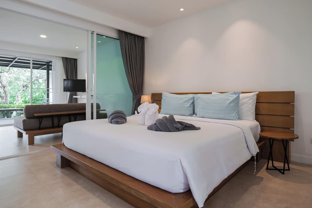 A modern hotel room with a large bed, a sofa, a TV, and a view of greenery.