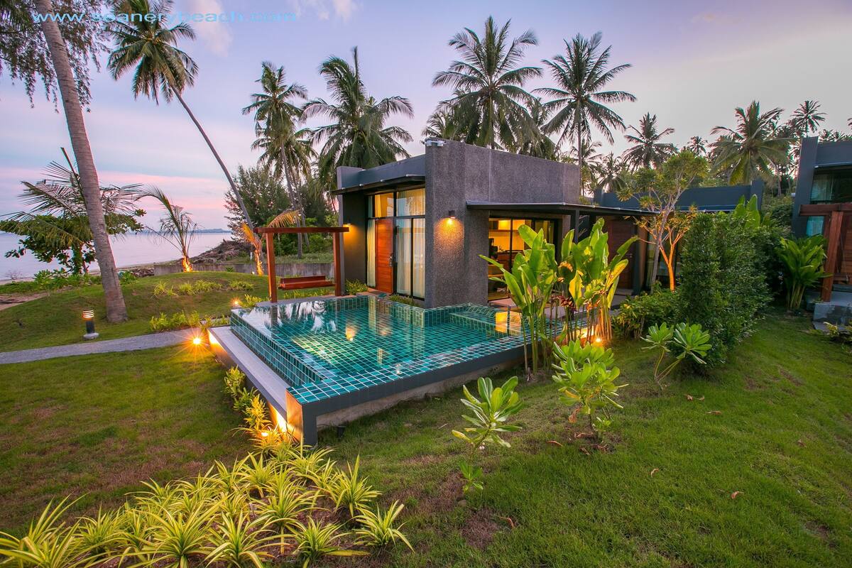 A modern villa with a glass infinity pool, surrounded by lush greenery and palm trees, overlooking a body of water.