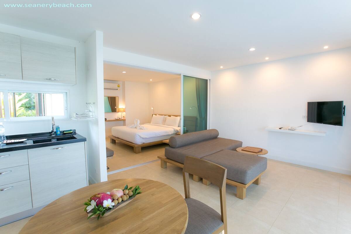 A modern hotel room with a kitchenette, a dining area with a wooden table, a sofa, and a TV mounted on the wall.