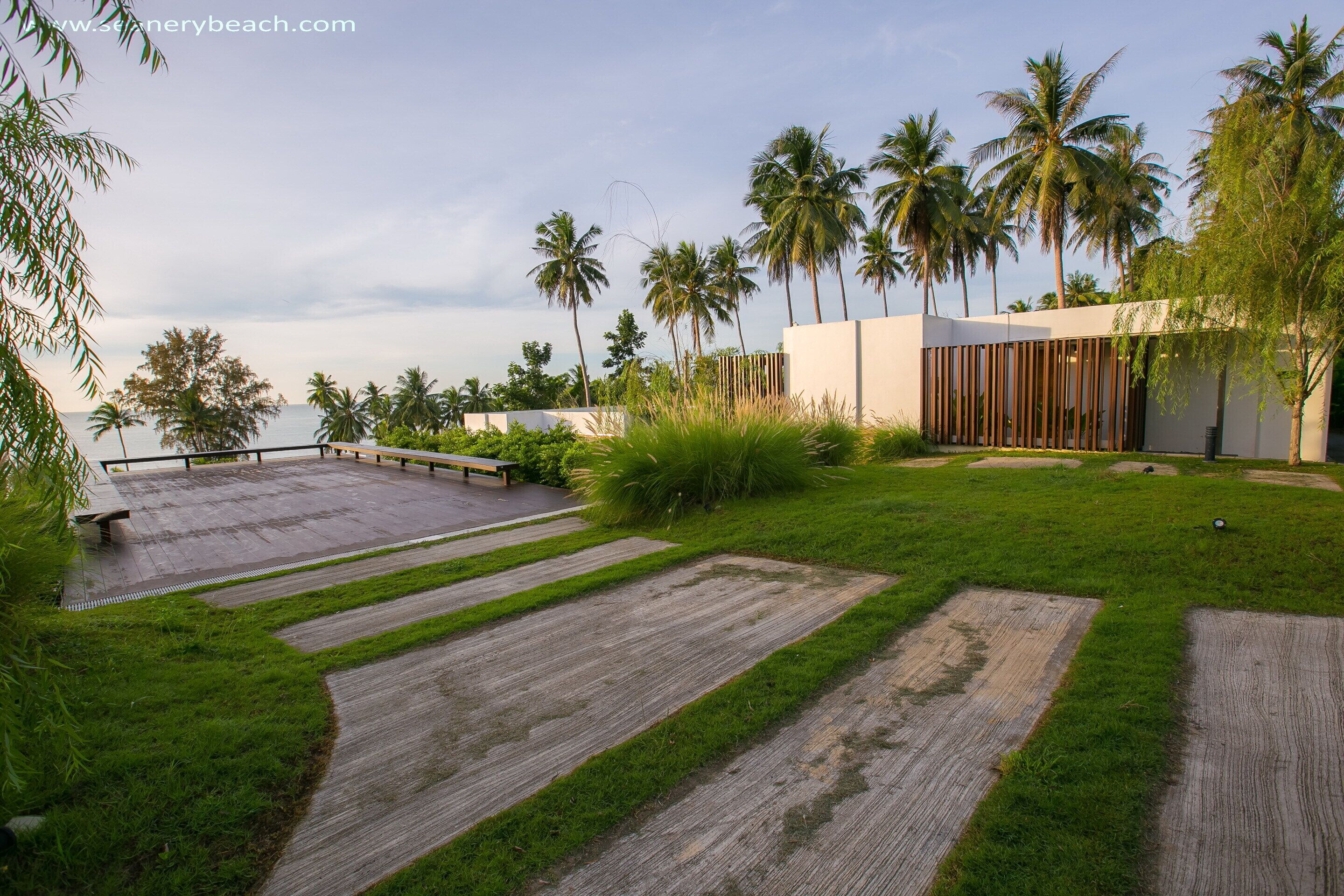 A modern outdoor area with a wooden deck, grassy patches, palm trees, and a white building with vertical slats.