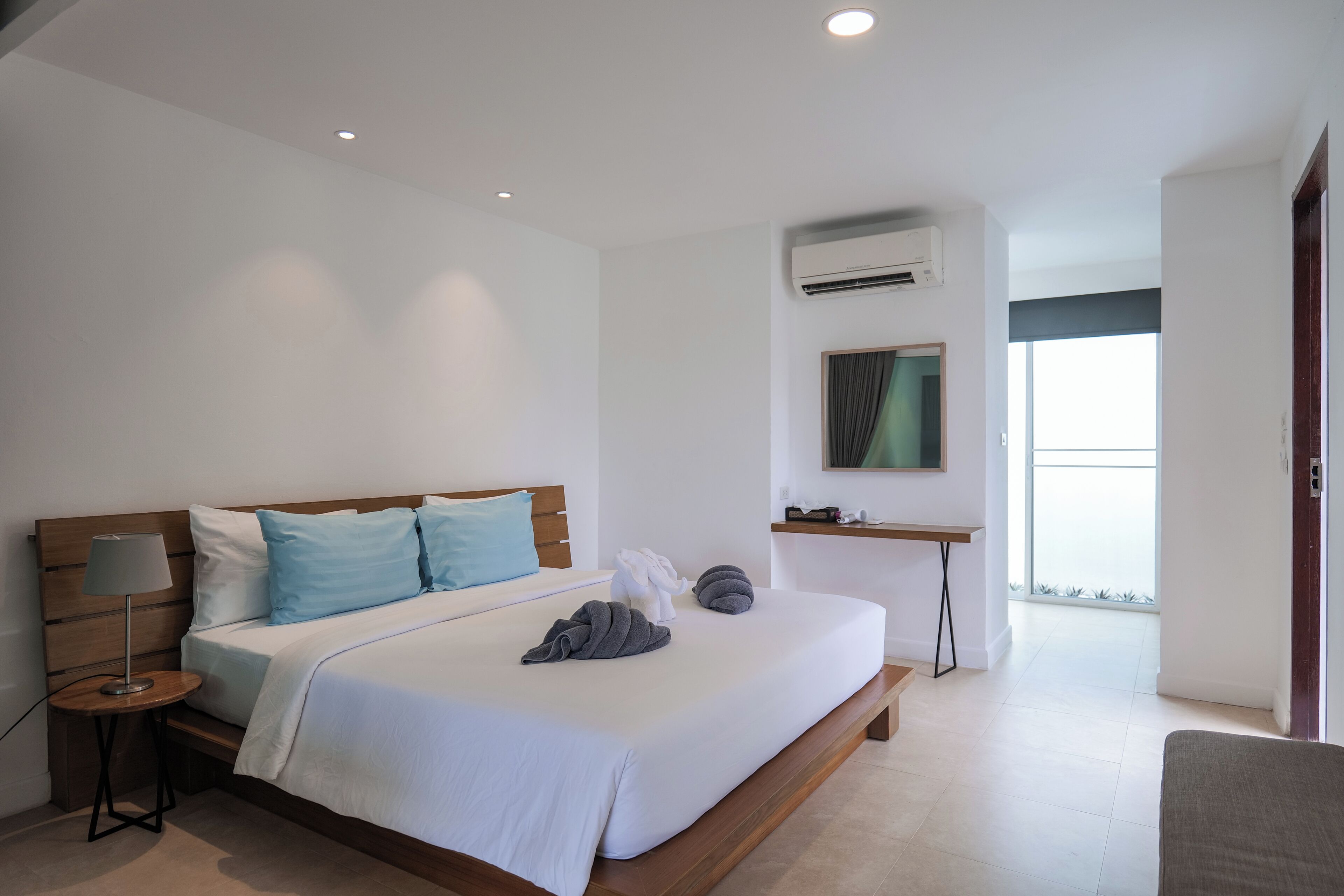 Habitación de hotel moderna con cama de madera, ropa de cama blanca, una pequeña mesita de noche con lámpara, un escritorio y un amplio ventanal con vistas al exterior.