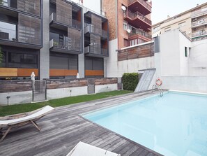 Un complejo de apartamentos moderno con piscina, terraza de madera y mobiliario exterior.