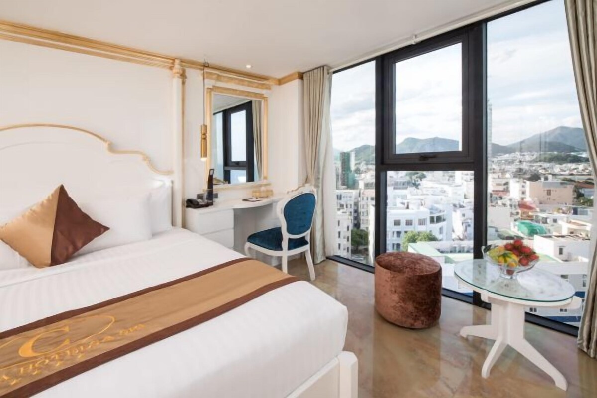 Ett hotellrum med en stor säng, ett skrivbord, en stol, ett litet bord med en vas med blommor och utsikt över en stadspanorama genom stora fönster.