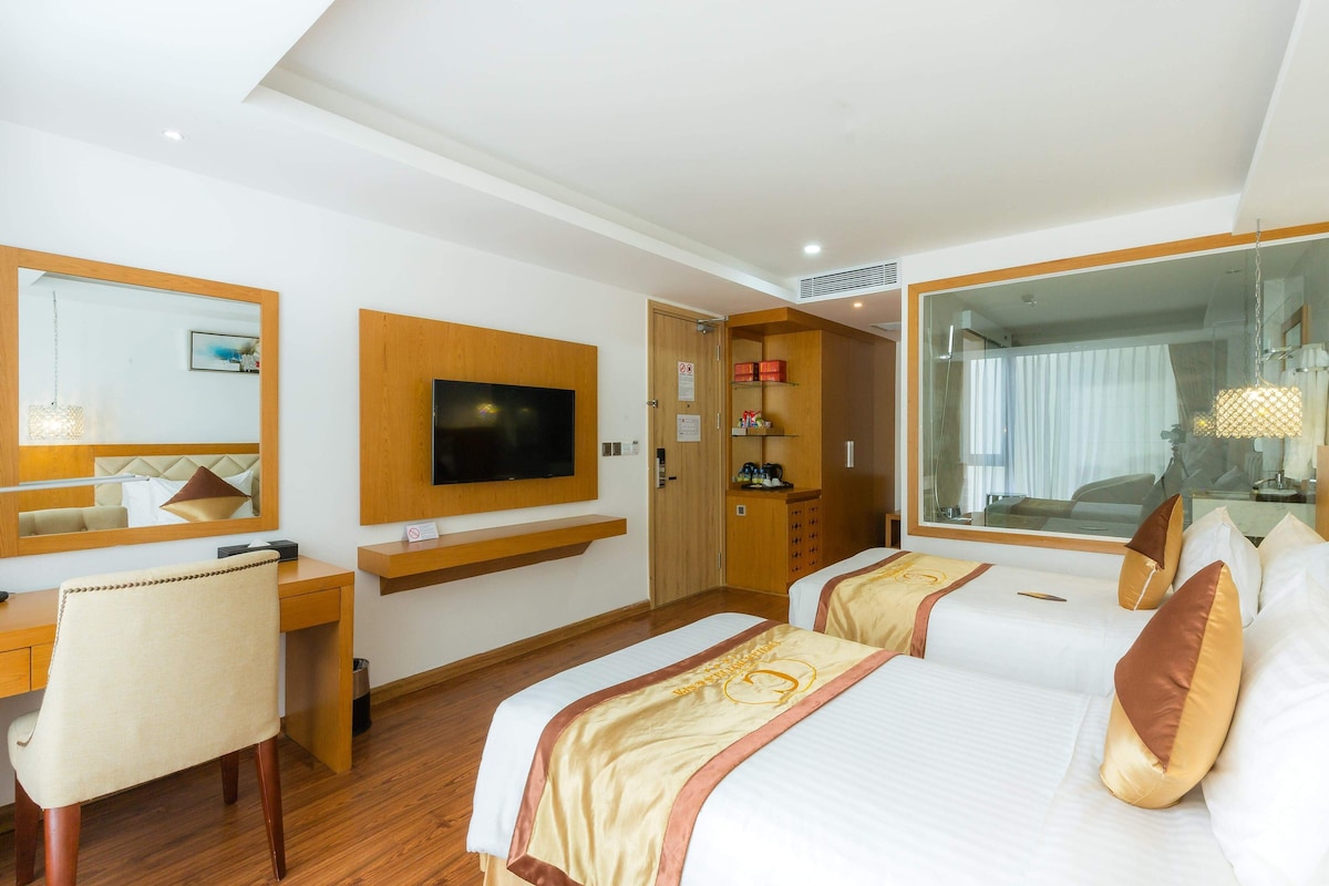 A hotel room with a desk, two beds, a TV, a mirror, and a large window.