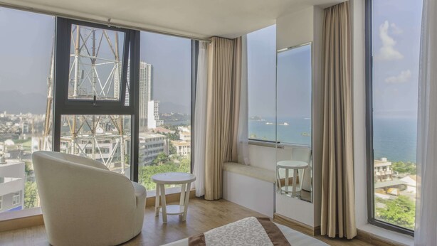 Una habitación moderna con ventanas amplias, un sillón curvo, una pequeña mesa redonda y vistas a la ciudad y al mar.