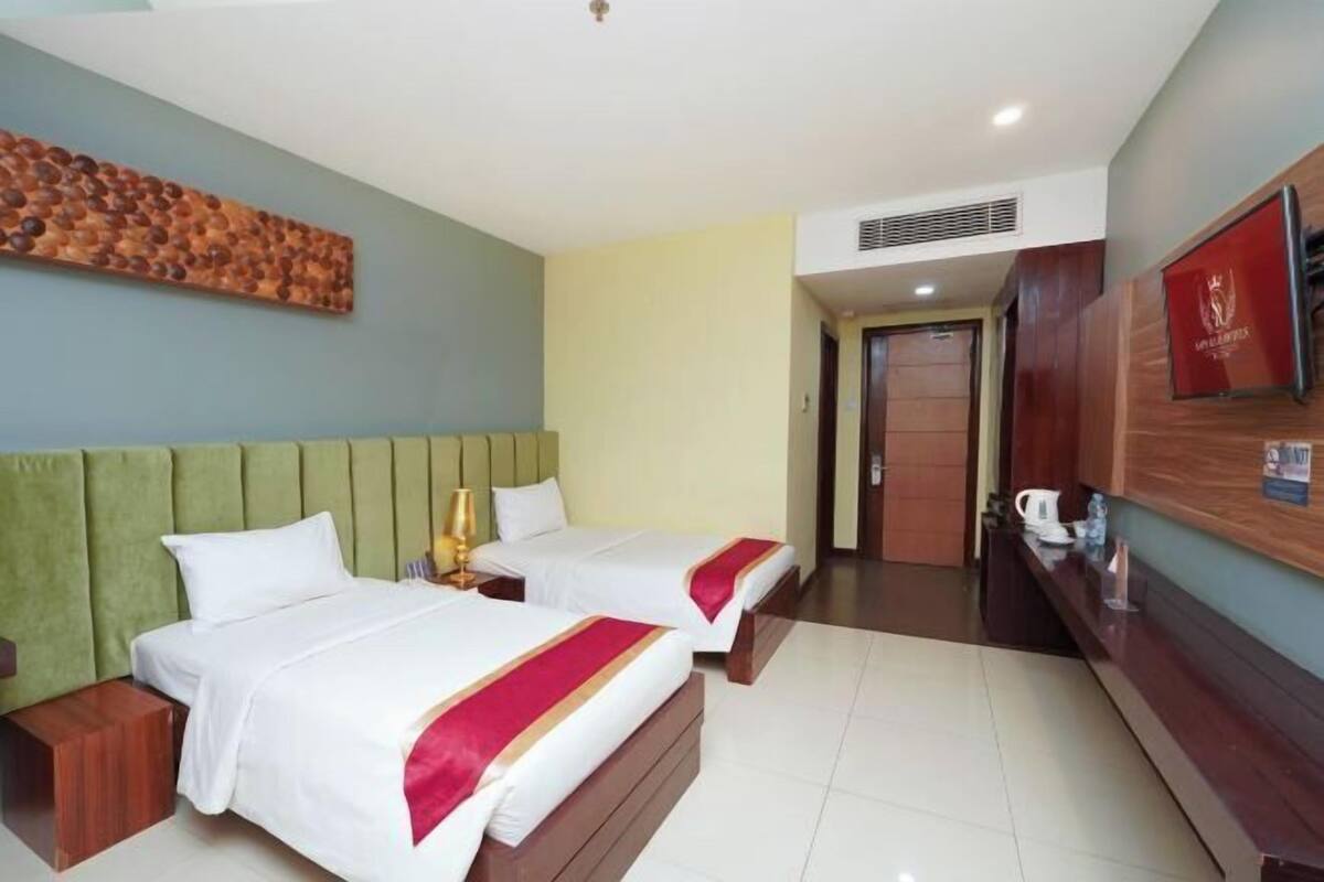 Une chambre d’hôtel avec deux lits, une tête de lit verte, une commode en bois, un téléviseur et une œuvre d’art accrochée au mur.