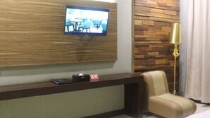 Een moderne hotelkamer met een houten accentmuur, een flatscreen televisie boven een donker console tafeltje, een gouden vloerlamp en een beige fauteuil.