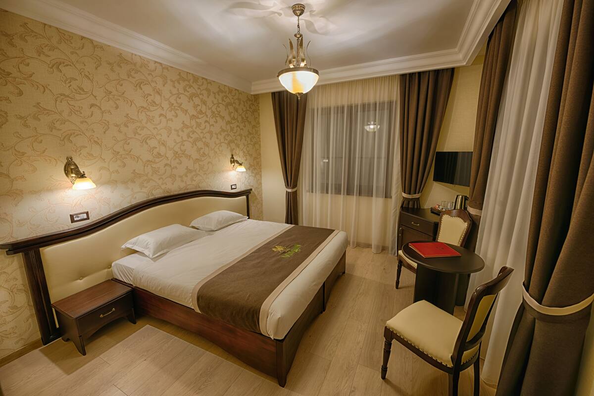 A hotel room with a large bed, bedside tables, a desk with a chair, and a window with curtains.