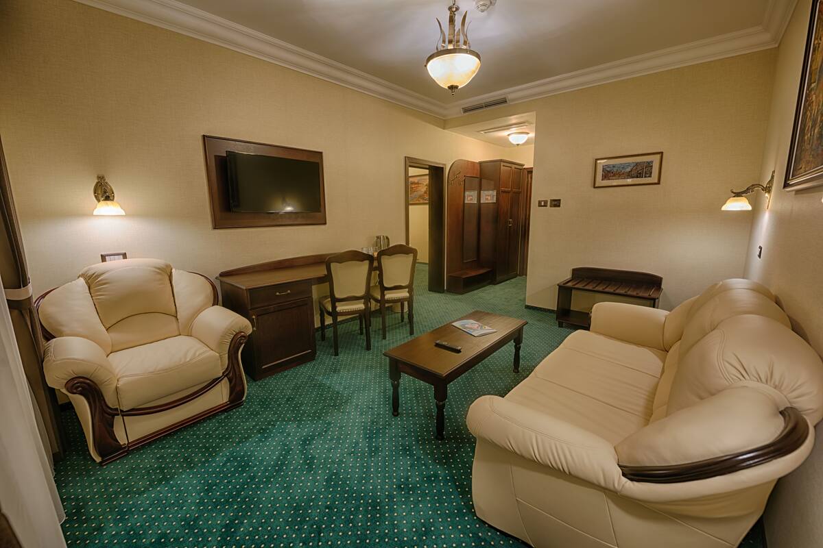 A hotel room with a green polka dot carpet, two beige leather armchairs, a wooden coffee table, a television mounted on the wall, and a wooden wardrobe.