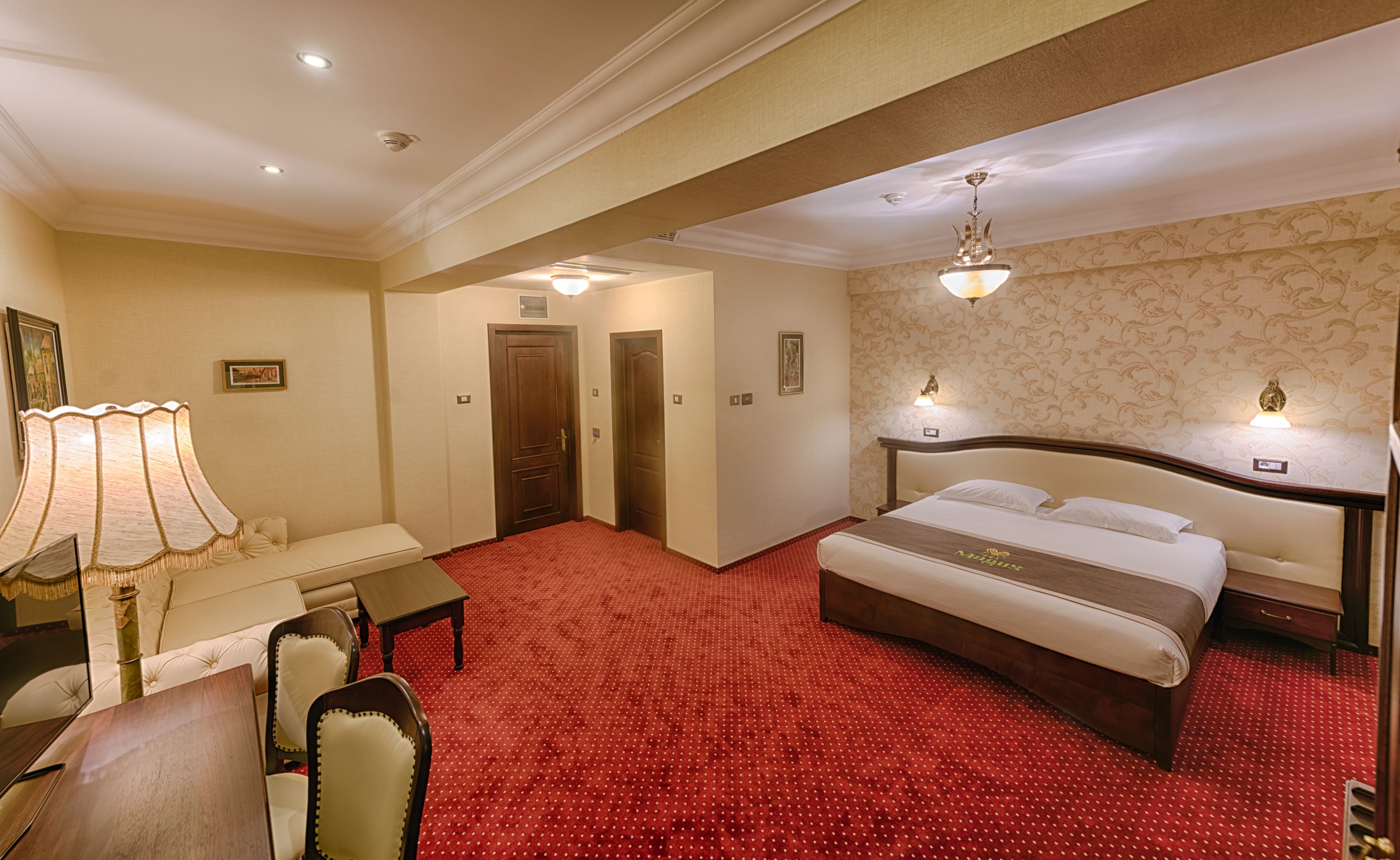 A hotel room with a large bed, a desk with a lamp, a chair, and a red carpeted floor.