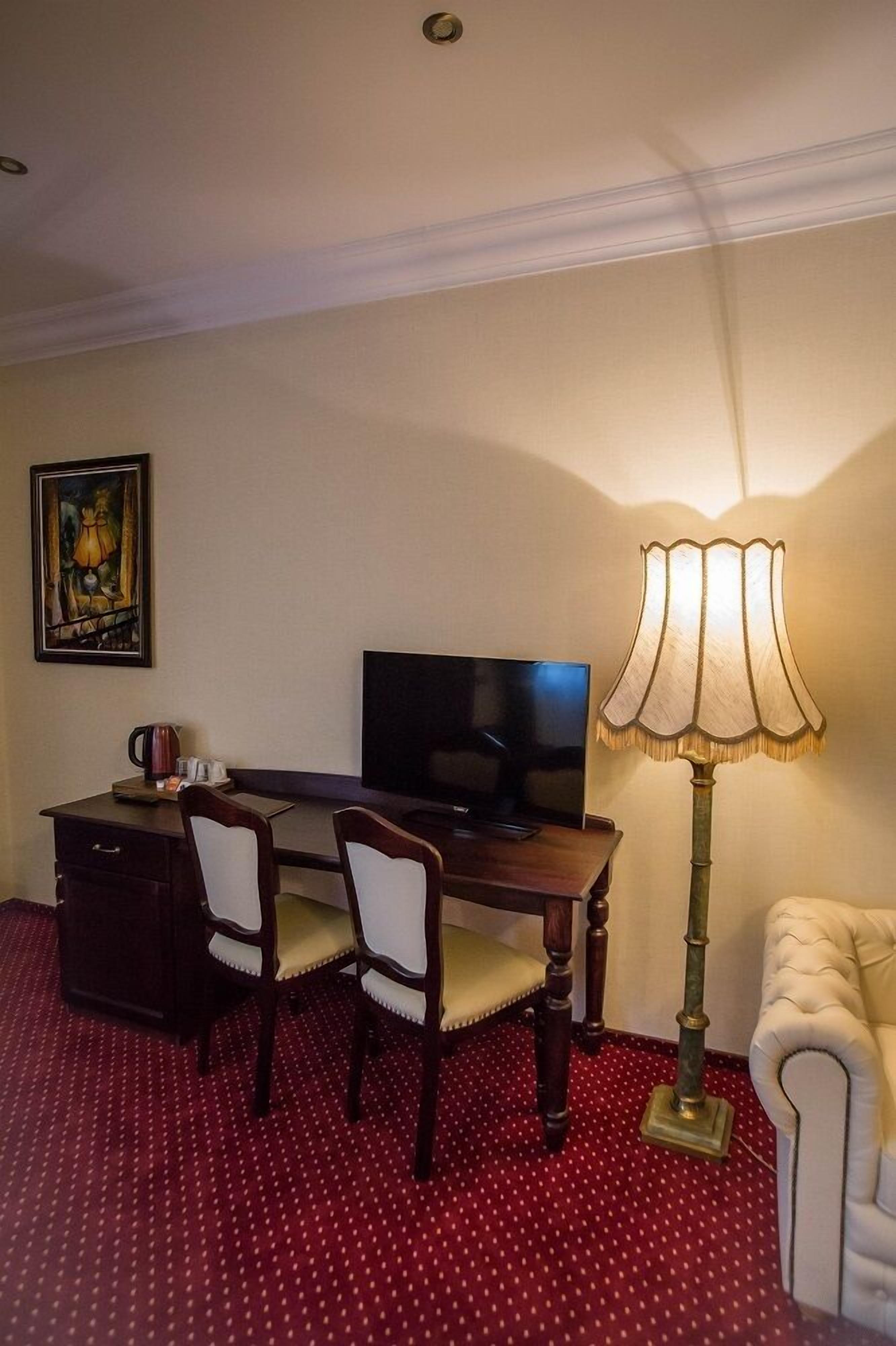 A hotel room with a desk, two chairs, a television, a lamp, a painting, and a red polka dot carpet.