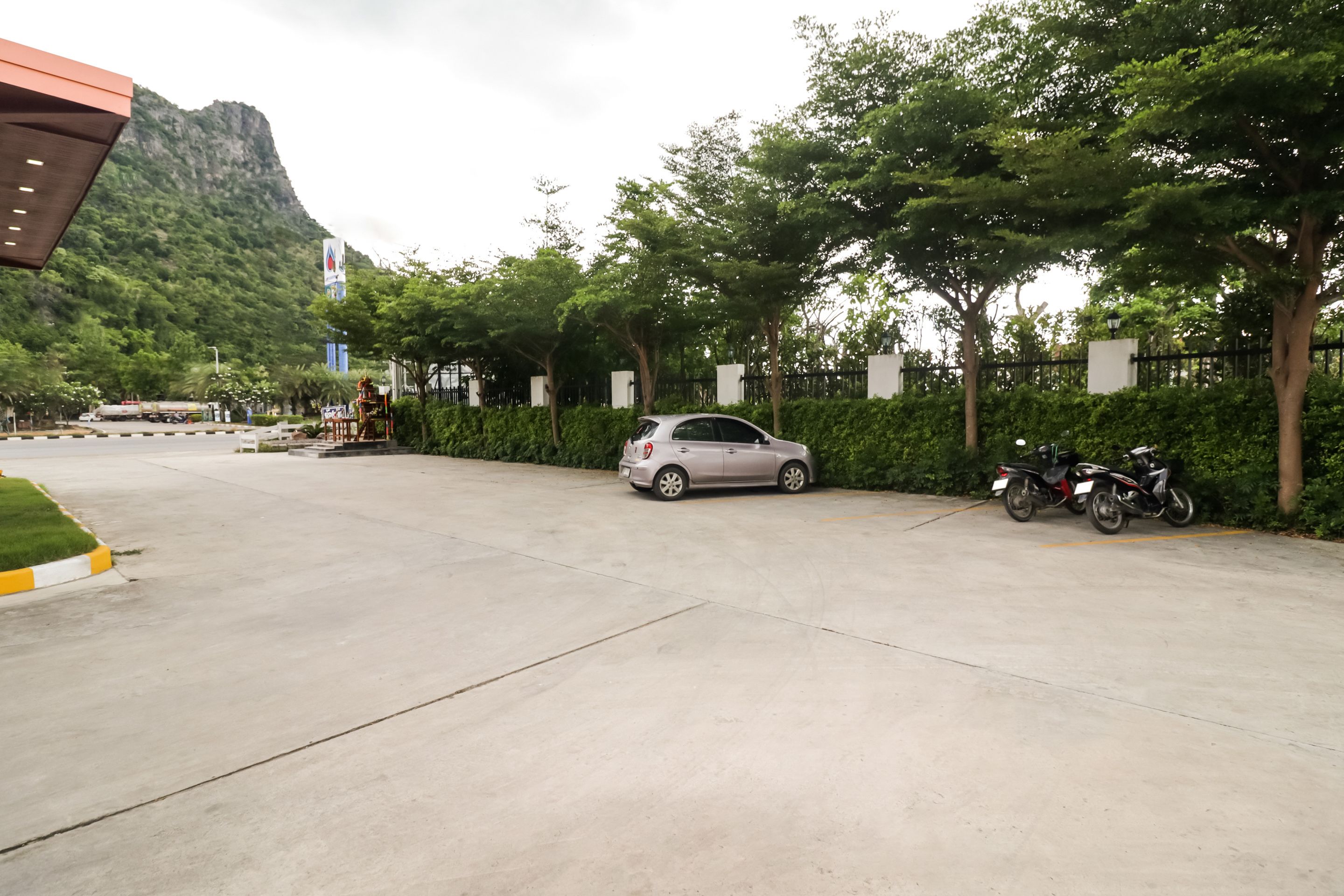 Un espace de stationnement où sont garées des voitures et des motos, entouré de verdure et avec une montagne en arrière-plan.