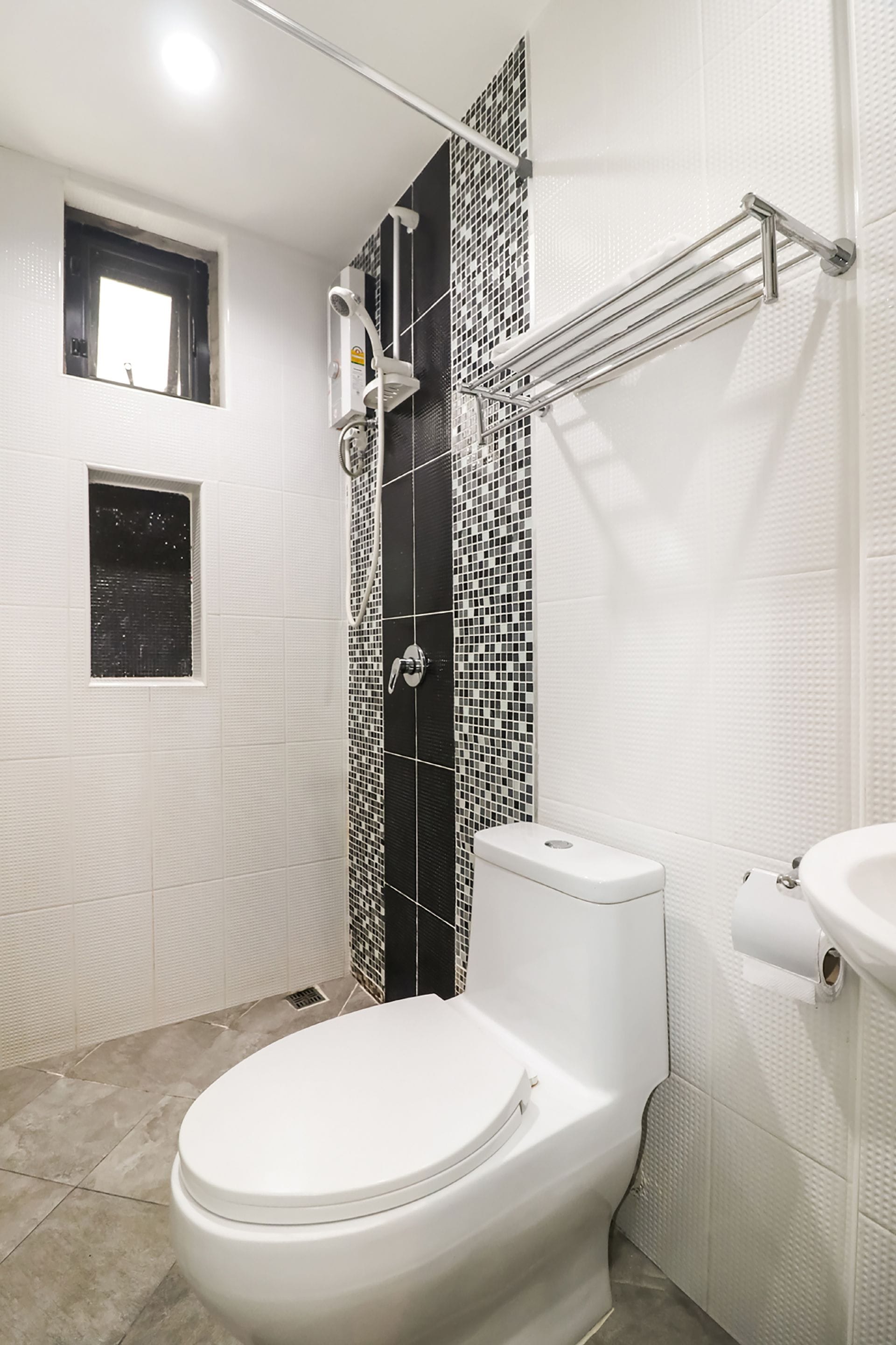 Un baño con un inodoro blanco, una zona de ducha con pared de azulejos blanco y negro y un ventanal pequeño.