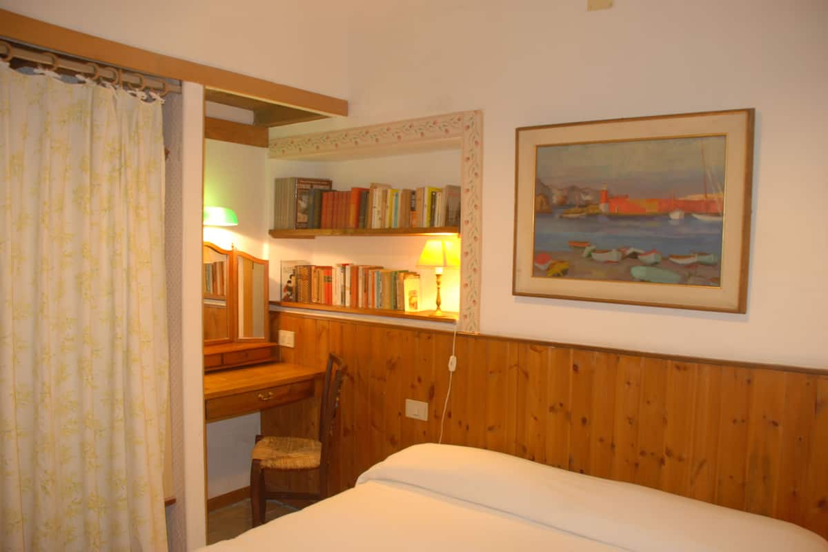 A room with a bed, a wooden desk, a chair, a bookshelf, and a painting of boats on the wall.