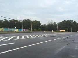Una intersección de calles con varios carriles y semáforos. Hay vallas publicitarias y señales a un lado de la vía.