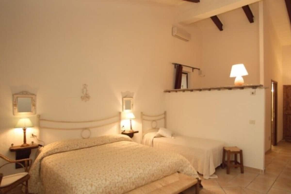 A bedroom with a large bed, two bedside tables with lamps, a small stool, and a wooden door.