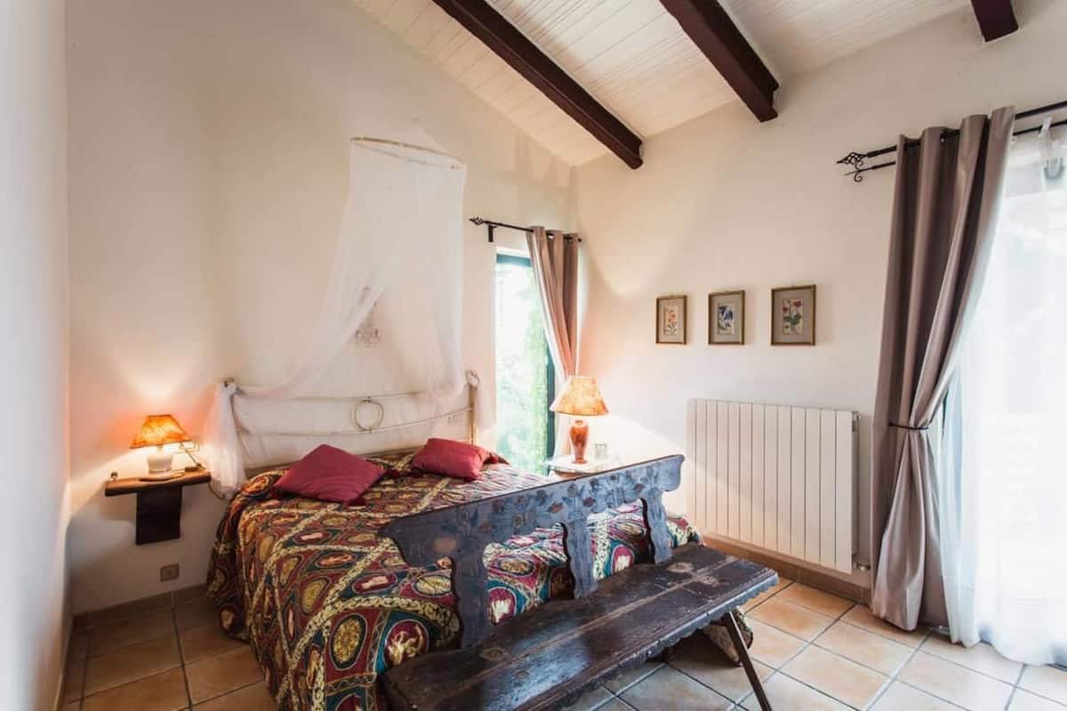 A bedroom with a bed, bedside lamps, a bench, a window with curtains, and a radiator.