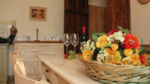 Una cocina con una mesa de madera, una cesta de mimbre con flores, copas de vino y una botella.