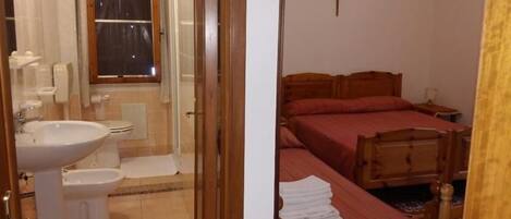 A bathroom with a sink, toilet, and shower. The room has a wooden bed with red bedding and a cross on the wall.
